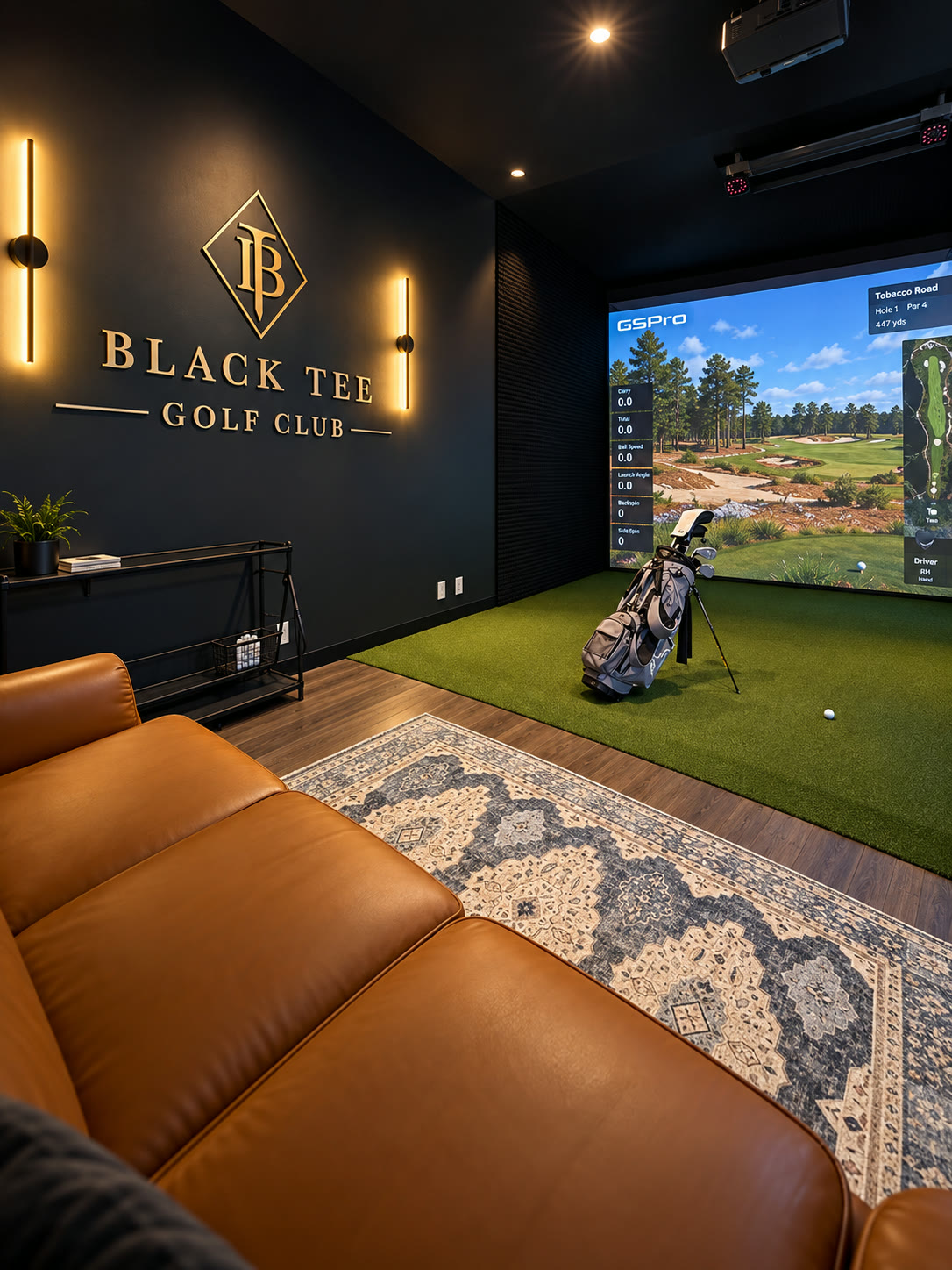 Leather sofa facing a Black Tee bay with a golf bag on the turf, GSPro on the screen and the gold Black Tee Golf Club sign