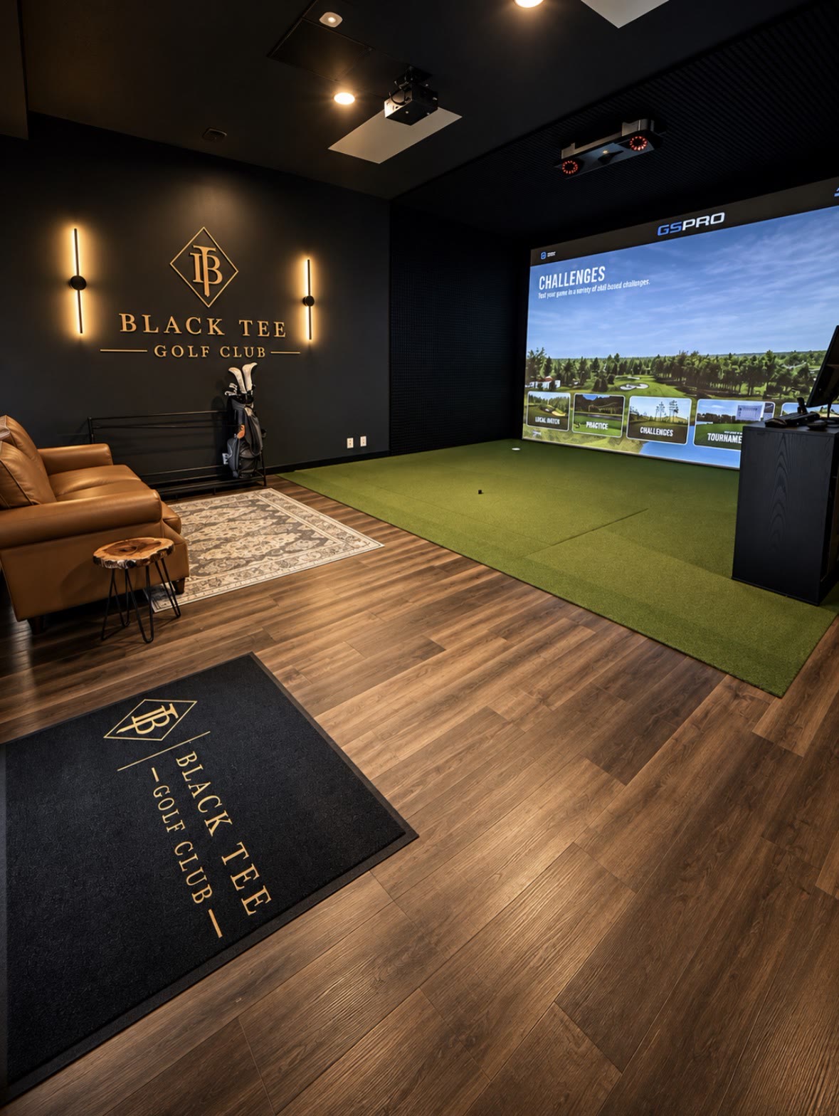 Black Tee simulator bay with GSPro challenges on the screen, gold Black Tee Golf Club wall sign, leather seating and branded floor mat